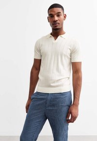 Polo à manches courtes, crème, avec une texture côtelée et des rayures verticales, associé à un pantalon rayé bleu et blanc.