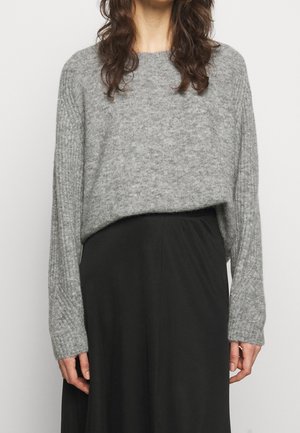 Femme portant un pull en maille côtelée gris à manches longues et une jupe noire taille haute, debout devant un fond uni.