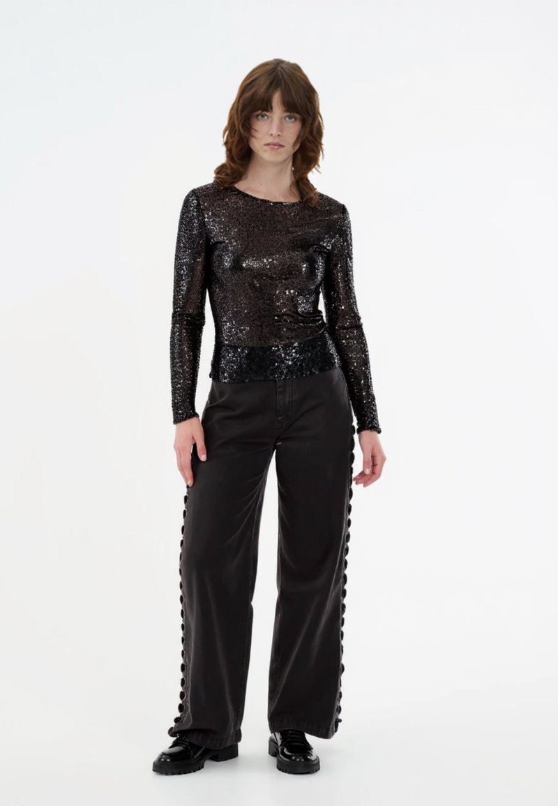 Μαύρο μακρυμάνικο τοπ με υφή από πούλιες, σ�υνδυασμένο με μαύρα παντελόνια με φαρδιά πόδια που έχουν κουμπιά κατά μήκος των πλευρικών ραφών.