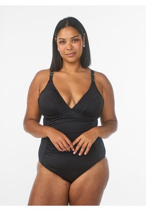 Donna con carnagione media che indossa un costume intero nero, in piedi con le mani intrecciate su sfondo bianco.