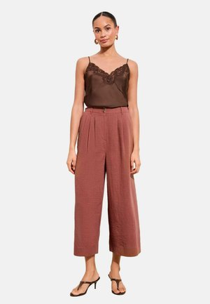 Žena stojaca na bielom pozadí, oblečená v hnedom sťahovacom topíku s čipkovaným lemom, tehlovo sfarbených širokých nohaviciach s dĺžkou nad členky, hnedých sandáloch na opätku a zlatých kruhových náušniciach.