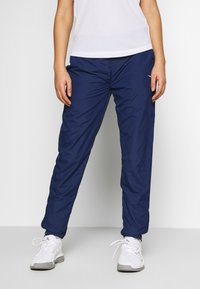 Námořnické modré sportovní kalhoty vyrobené z lehkého materiálu, s tapered střihem a zipy u kotníků; kombinované s bílými sportovními botami.