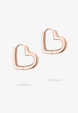Guldøreringe i hoop med et geometrisk design, der har en poleret overflade. Varme hudtoner og neutral makeup komplimenterer en mørk mønstret blazer.