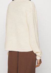 Cream-colored knitted sweater with a loose fit, ribbed cuffs and hem, textured fabric, worn over brown pants.