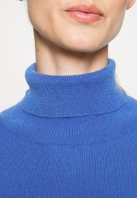 Niebieski sweter z golfem o prążkowanej fakturze. Kołnierz jest dopasowany, a materiał wydaje się miękki i dziany, podkreślając czysty design.