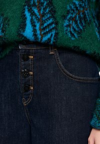 Dark blue denim jeans featuring five buttons, contrasting stitching, and a front pocket, partially covered by a green and blue patterned sweater.