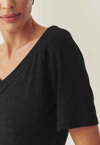 Čierny krátky rukáv s hlbokým výstrihom do V a jemným, textúrovaným povrchom. Tkanina má jemný lesk, ktorý pridáva hĺbku pevnej farbe.