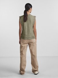 Pitkähiuksinen nainen, jolla on tummat hiukset poninhännällä, pukeutuneena vihreään pitsiseen hihattomaan toppiin ja löysiin beigenvärisiin housuihin, seisoo yksinkertaisen vaalean taustan edessä.