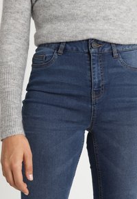 Jeans a vita alta di colore blu scuro con una texture liscia, design classico a cinque tasche e cuciture arancioni a contrasto. Chiusura con bottone e zip.