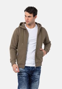 Olijfgroen zip-up hoodie met een trekkoordkap, voorzakken en geribbelde manchetten, gedragen over een witte t-shirt en gecombineerd met denim jeans.
