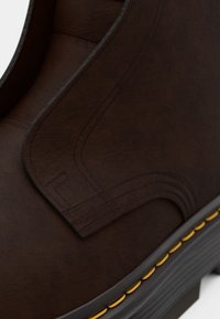 Bottine en cuir marron avec des coutures courbées, une surface texturée et des accents jaunes le long de la semelle. Présente un design arrondi.
