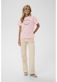 Smaidoša sieviete beigu biksēs un rozā T-kreklā ar tenisa ilustrāciju, stāvot ar vienu roku aiz muguras pret vienkāršu fona.