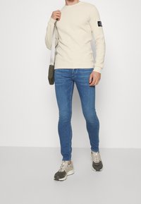 Man i beige långärmad skjorta, blå smala jeans och beige sneakers som håller i remmen till en olivgrön axelväska.