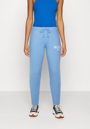 Person iført lyseblå joggingbukser med "Hollister" logo og sorte og hvide sneakers, der står mod en ensfarvet baggrund.