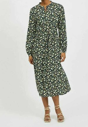 Kobieta ubrana w czarną sukienkę midi z długim rękawem, ozdobioną drobnym białym i żółtym motywem kwiatowym, oraz beżowe sandały na paski.