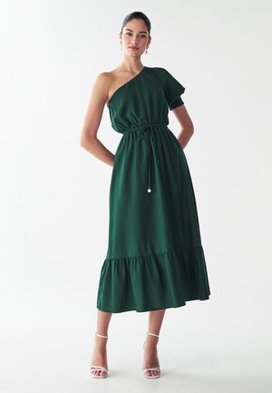 Groene een-schouderjurk met een gathered taille, textuurstof en een uitlopende rufflezoom. Draagt men met witte gesp praktische hakken.