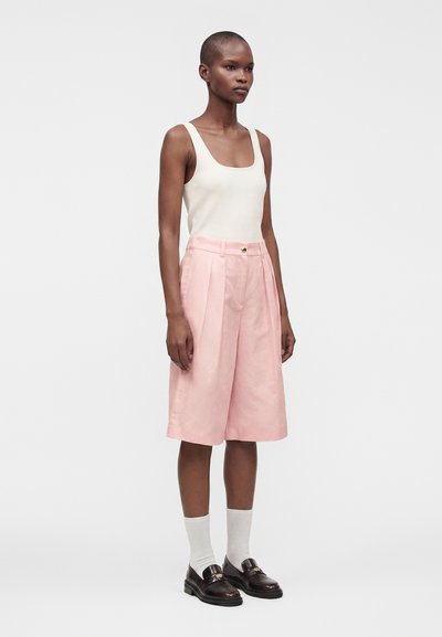 Femme debout portant un haut blanc sans manches, un short rose taille haute arrivant au genou, des chaussettes blanches et des mocassins noirs.