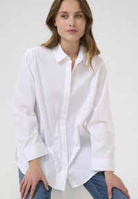 Camicia bianca a maniche lunghe con colletto classico, vestibilità rilassata e texture liscia, abbinata a jeans in denim blu.