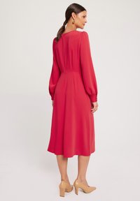 Czerwona sukienka z długim rękawem, o plisowanym wzorze, okrągłym dekolcie i guzikami przy mankietach, zestawiona z cielistymi szpilkami.
