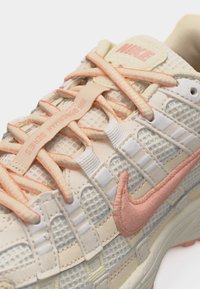 Beige and peach Nike sneaker with mesh upper, peach embroidered swoosh, and peach-accented laces threaded through fabric loops.
