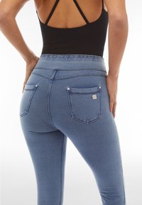 Nainen, joka pukeutunut korkeavyötäröisiin sinisiin farkkuhousuihin ja mustaan remmipaitaan, kuvattuna takaa käsien ollessa lanteilla.