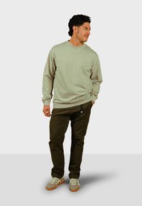 Lichtgroene sweatshirt met geribbelde boorden, gecombineerd met donkergroene broek met een accent van een voorvak. Casual schoenen in grijs en bruin.
