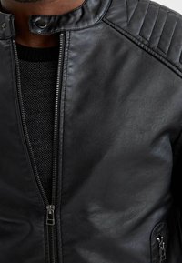 Veste en cuir noir avec un col arrondi, des épaules matelassées et une fermeture éclair à l'avant avec des éléments métalliques. Doublure texturée visible.