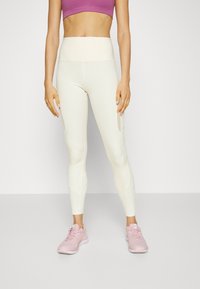 Leggings de cintura alta en crema con paneles laterales texturizados, combinados con una camiseta corta morada y zapatillas deportivas rosas claras. Diseño liso y ajustado.