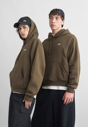 To unge voksne som har på seg olivengrønne Nike-hetter og mørke bukser, den ene med hetta på og svart caps, den andre med blå caps på bakvendt.