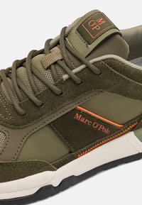 Oliivinvihreät ja beige tennarit, joissa on samettisia ja verkkopaneeleja, oranssia ompelua ja brändäystä, valkoinen välikerros ja musta kumipohja.