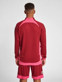 Mörk röd sportjacka med rosa detaljer, långa ärmar och hög krage. Har nätpaneler på axlarna och matchande mörkröda shorts.