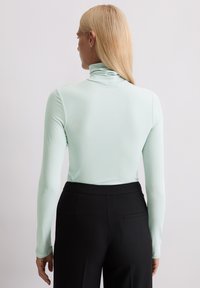 Helles mintgrünes Langarm-Turtleneck-Oberteil mit weicher Textur, tailliertem Design und hohem Kragen, kombiniert mit schwarzen maßgeschneiderten Hosen.