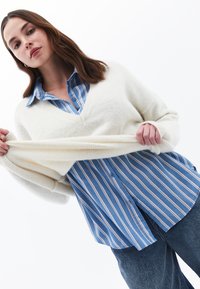 Maglione bianco, morbido e strutturato con scollatura a V indossato sopra una camicia azzurra a righe con bottoni. Il maglione ha maniche ampie e polsini a coste.