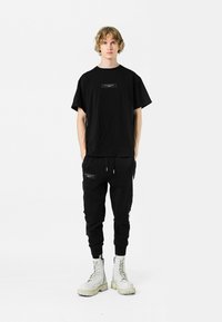 Camiseta de algodón negra con un logo rectangular en el pecho, combinada con joggers negros con bolsillos con cremallera y botas de combate con suela blanca.