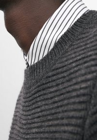 Suéter gris de punto con una textura acanalada, colocado sobre una camisa blanca de cuello con finas rayas verticales negras.