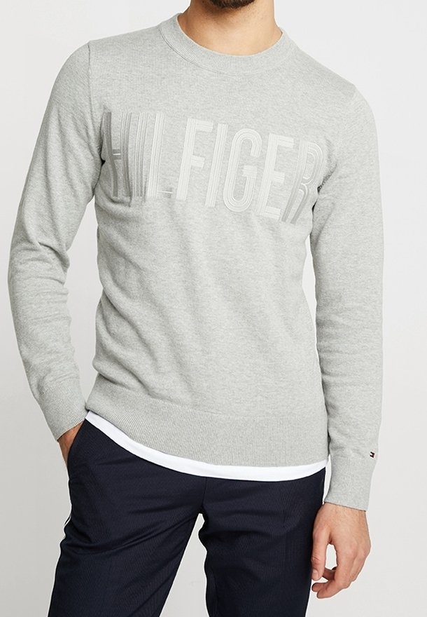 Homme portant un pull léger gris clair col rond avec le texte « HILFIGER » sur la poitrine et un pantalon foncé, debout avec une main dans la poche.