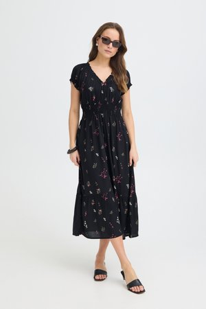 Kobieta w czarnej sukience midi w kwiaty, czarnych sandałach typu klapki, okularach przeciwsłonecznych i czarnej bransoletce, stojąca na jednolitym jasnym tle.