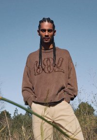 Brun stickad tröja med "Cool" i texturerad lettering, tillsammans med beiga byxor. Tröjan har en avslappnad passform och ribbade ärmslut.