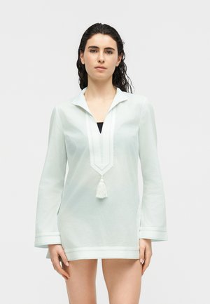 Tânără cu păr închis la culoare poartă o tunică albă transparentă cu mâneci lungi, decolteu adânc în V și detalii cu ciucuri, stând în fața unui fundal simplu.