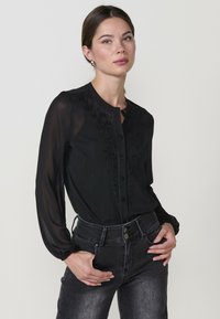 Juoda ilgomis rankovėmis bliuska su siuvinėjimu, mygtukų užsegimas priekyje ir šiek tiek rauktomis rankovėmis. Suderinta su tamsiomis džinsinėmis kelnėmis.