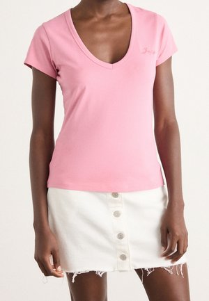 Femme portant un t-shirt rose clair à manches courtes et col en V ainsi qu'une mini-jupe en jean blanche à boutons sur le devant avec un ourlet effiloché.
