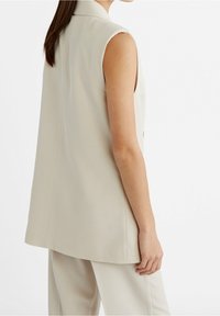 Kobieta w bezrękawnikowej beżowej kamizelce dopasowanej do sylwetki i pasujących spodniach, widoczna od tyłu na jednolitym białym tle.