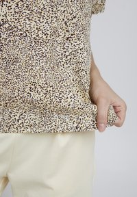 Top v leopardím vzoru v béžové a hnědé barvě, krátké rukávy, kulatý výstřih, měkká tkanina, kombinovaný se světle žlutými kalhotami, ruka držící lem.