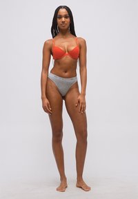 Γυναίκα με μακριά πλεξούδα, ξυπόλυτη, φοράει πορτοκαλί σουτιέν και γκρι σλιπ Emporio Armani σε απλό φόντο.