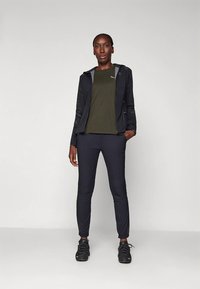 Crna atletska jakna laganog dizajna, maslinasto zelena košulja i mornarsko plave hlače. Crne tenisice upotpunjuju odjevnu kombinaciju.