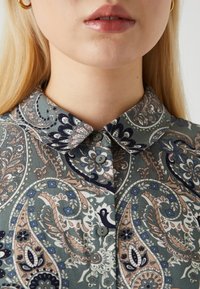 Button-up shirt in green fabric with paisley pattern. Features a collared neckline, light buttons, and intricate floral designs in pink and blue.
