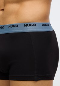 Boxers pretos com uma cintura elástica azul claro decorada com o logótipo preto "HUGO". O tecido parece suave e elástico.