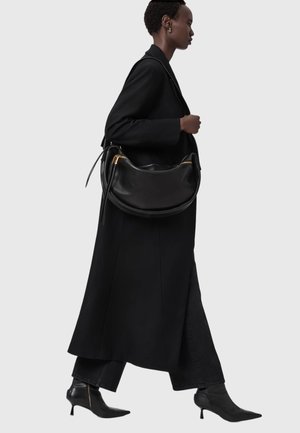 Zwarte leren schoudertas met een gebogen vorm, gouden metalen details en een gladde textuur. Gedragen aan de zijkant tegen een zwarte jas.