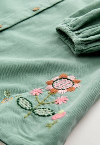Grüne Cordhemd mit floraler Stickerei in Pink und Orange, mit Knöpfen und einem gerafften Bündchen. Strukturierter Stoff mit kunstvollen Details.