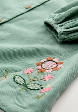 Grön kordflor-shirt med blommig broderi i rosa och orange, med knappar och en volangärms. Texturerad tyg med invecklade detaljer.
